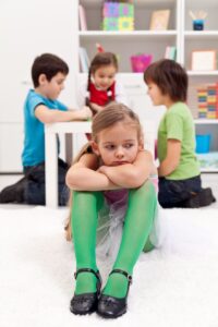 young girl in a classroom sitting alone and looking sad as children play together behind her