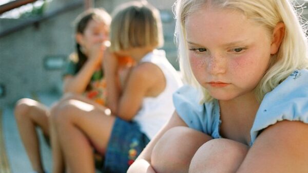 Sad blond girl sitting alone as girls talk and whisper behind her