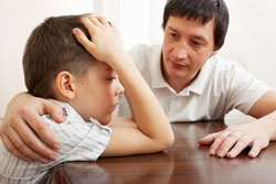 young boy at a table looking disappointed, being comforted by father