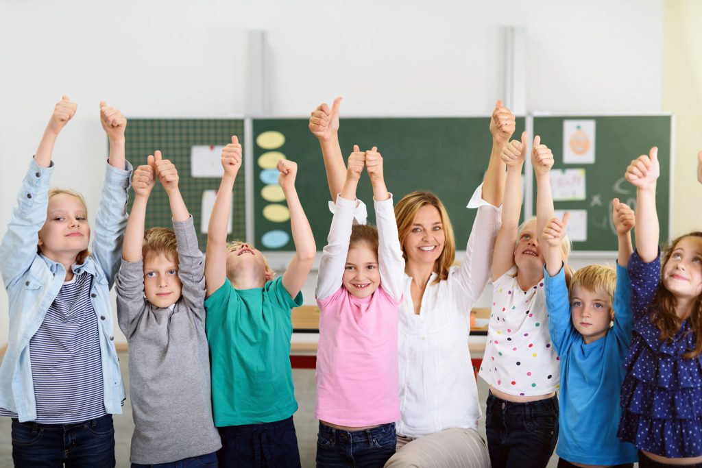 Happy teacher and children in classroom with thumbs up overhead